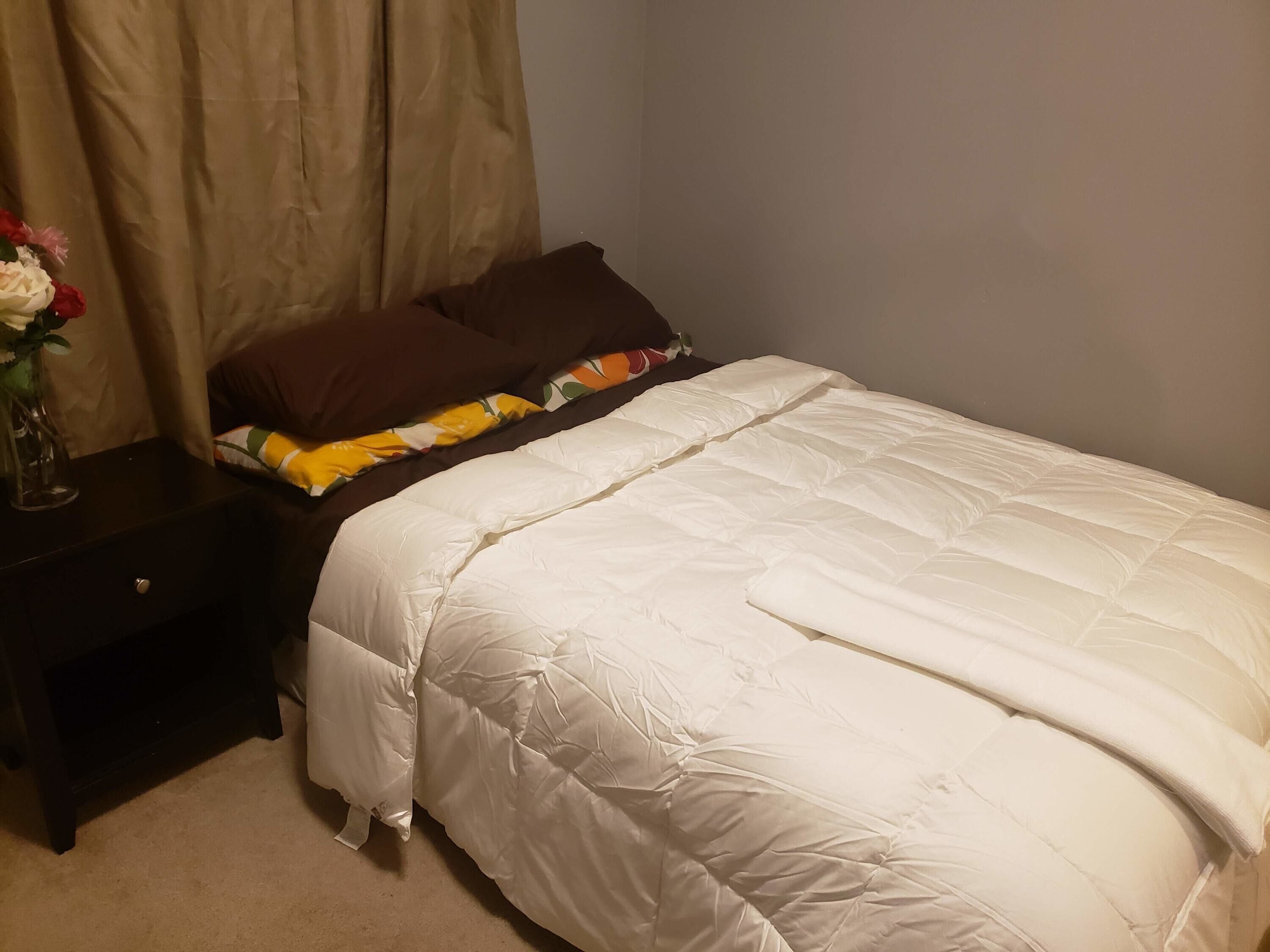 A neatly made bed with a white duvet and brown pillows, a bedside table with a vase of flowers, and a curtain partially drawn.