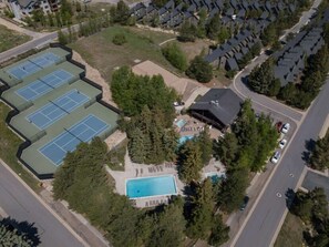An aerial view of a residential area with a swimming pool, tennis courts, and a building with a dark roof.
