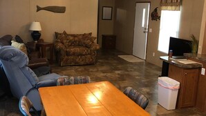 A living room with a wooden dining table, blue armchairs, a patterned sofa, a ceiling fan, and a fish decoration on the wall.