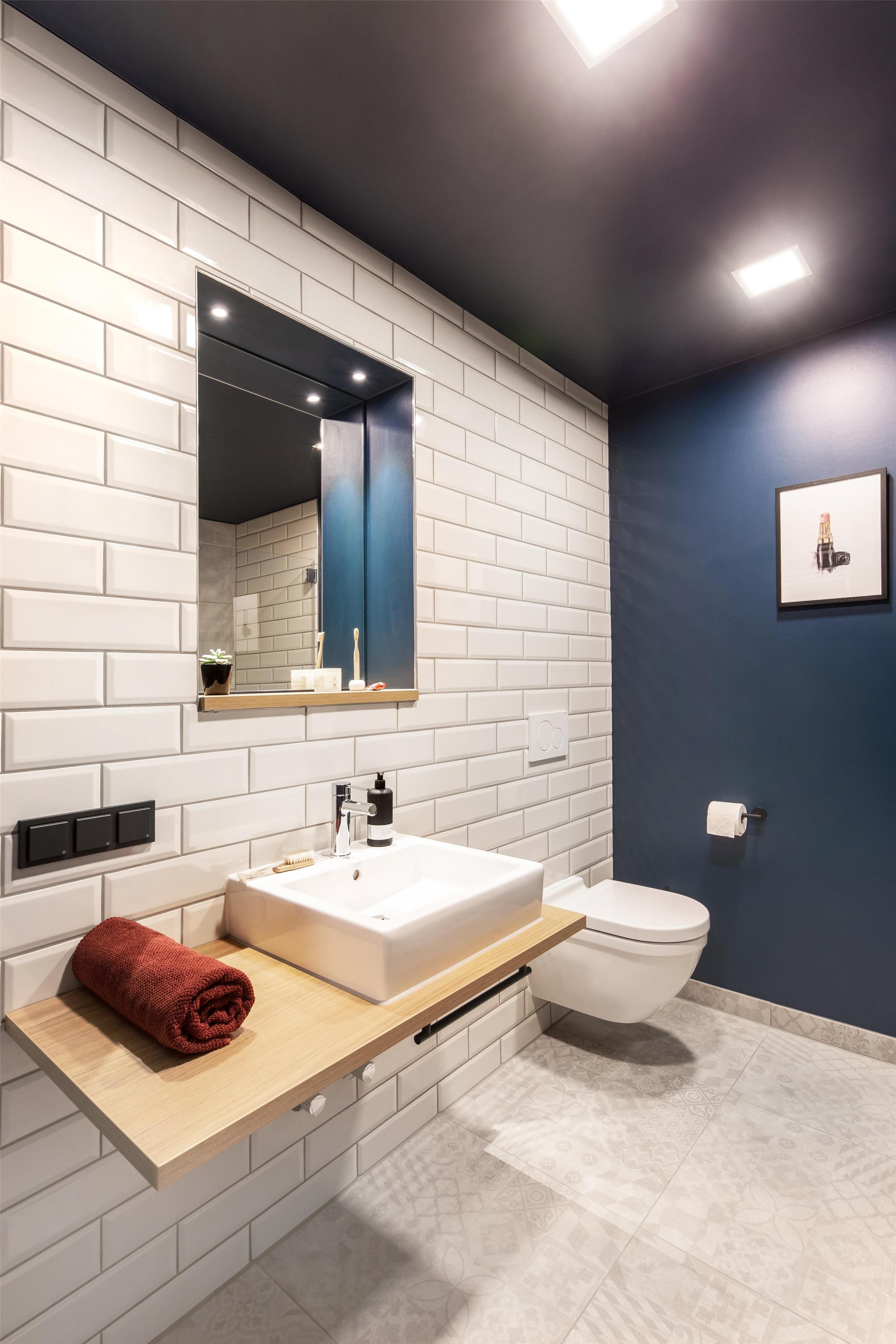 A modern bathroom with a white subway tile wall, a wooden vanity, a white sink, a mirror, a toilet, and a framed picture on the wall.
