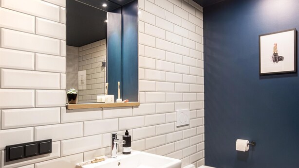 Un baño moderno con pared de azulejos blancos tipo metro, tocador de madera, lavamanos blanco, espejo, inodoro y un cuadro en la pared.