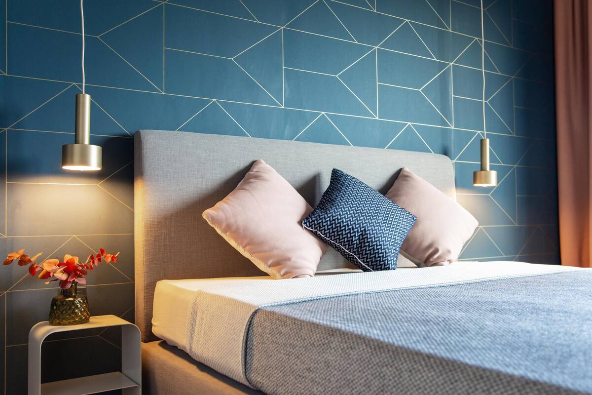 Une chambre à coucher moderne avec un mur orné d’un motif géométrique, une tête de lit rembourrée de couleur grise et deux lampes de chevet.