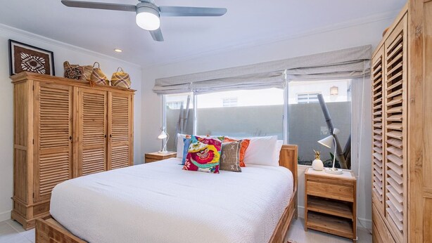 A bedroom with a bed, wooden wardrobe, ceiling fan, bedside lamps, and a window with curtains. - Caribbean Oasis - 3 spacious suites, private pool (Noord)