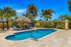 A swimming pool with lounge chairs and a thatched-roof structure, surrounded by palm trees and greenery.