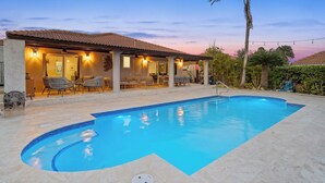 A swimming pool with a tiled deck, a covered patio with chairs, and a palm tree. - Caribbean Oasis - 3 spacious suites, private pool (Noord)