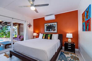 A bedroom with a bed, a ceiling fan, an air conditioning unit, a framed picture, and a small table with a lamp.
