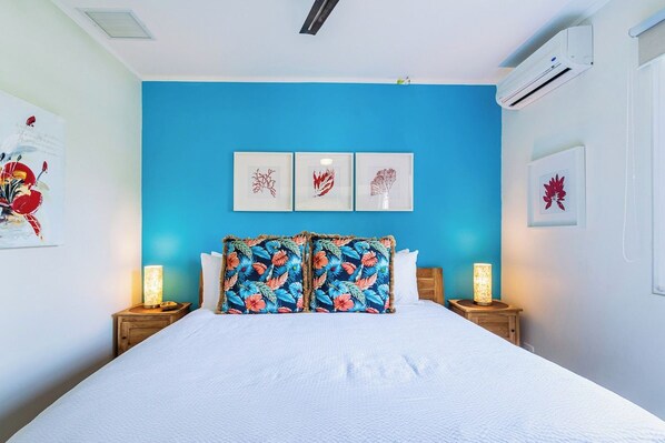 A bedroom with a blue accent wall, a bed with white bedding, two bedside tables with lamps, and three framed artworks above the bed.