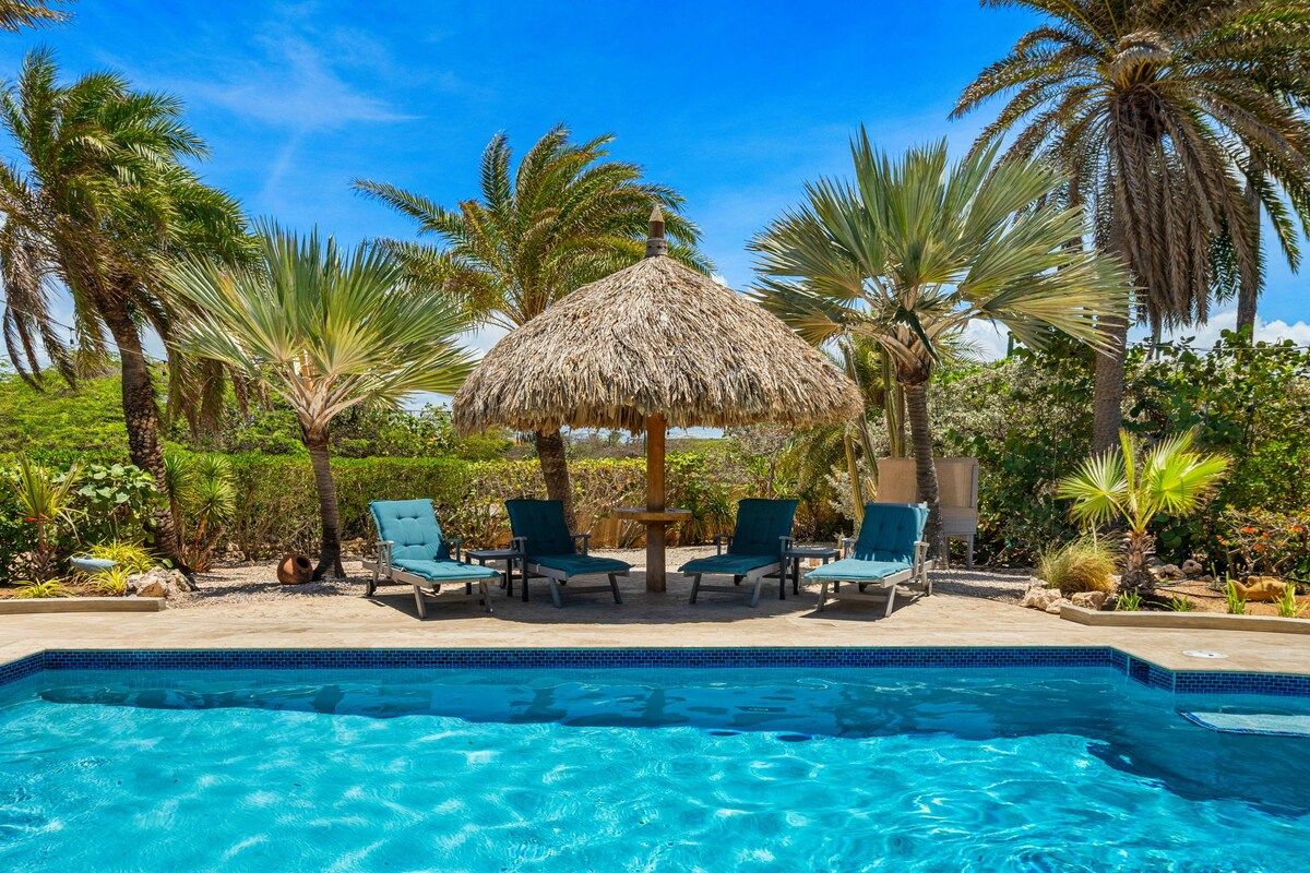 Une piscine entourée de chaises longues et d’un parasol en chaume, le tout bordé de palmiers et de verdure.