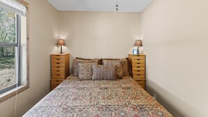 Un dormitorio con una colcha estampada, dos mesitas de noche de madera con lámparas, un ventilador de techo y una ventana con vistas a árboles.