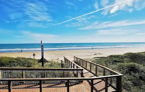 Une plage avec une promenade en bois menant au rivage, un mât de drapeau et un ciel dégagé.