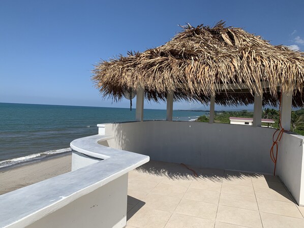 Une terrasse au bord de la plage, dotée d’une structure à toit de chaume, d’un banc blanc incurvé et offrant une vue sur la mer et les bâtiments situés au loin.