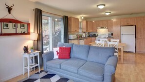 Ein modernes Wohnzimmer mit einem grauen Sofa, einem kleinen weißen Tisch mit einer Lampe sowie einer Küchenzeile mit Holzschränken und einem Kühlschrank.