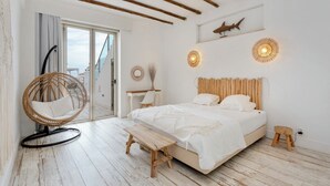 Une chambre à coucher comprenant un lit, un banc en bois, une chaise en osier, une petite table et un luminaire fixé au mur.