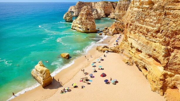 Une plage côtière bordée de falaises rocheuses, aux eaux turquoise et limpides, et d’un rivage sablonneux parsemé de parasols et de quelques personnes.