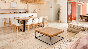 Une cuisine moderne avec des meubles blancs, un coin repas avec une table en bois et des chaises blanches, et un salon agrémenté d’un tapis à motifs et d’un canapé.