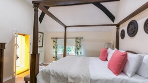 Un lit à baldaquin avec un couvre-lit blanc et des coussins roses, un ventilateur de plafond en bois et une fenêtre ornée de rideaux à motifs floraux.