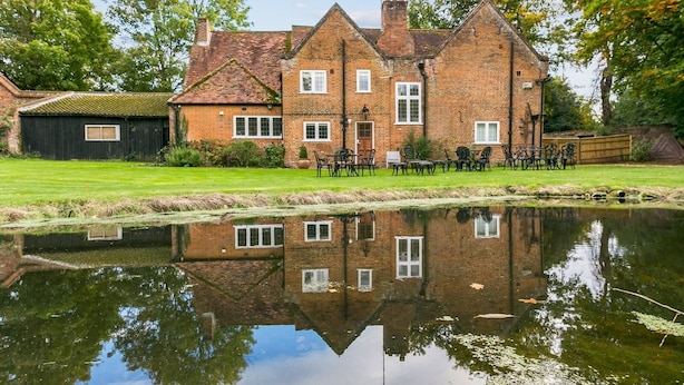 Une maison traditionnelle en briques, avec un étang devant, entourée de verdure et d’une clôture en bois.