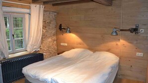 Un dormitorio de cabaña de madera con una cama, pared de piedra, vigas de techo de madera y dos lámparas fijadas a la pared.