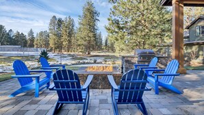 An outdoor patio with blue Adirondack chairs, a stone fire pit, and a stone grill. - Deschutes Executive Retreat - 6BR Luxury Rental (Deschutes County)