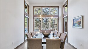 A dining room with a long table, chairs, a chandelier, and large windows with a view of trees. - Deschutes Executive Retreat - 6BR Luxury Rental (Deschutes County)