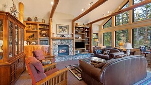 A cozy living room with a stone fireplace, wooden shelves, and a large window offering a view of the outdoors. - Rustic Ski-In/Ski-Out Townhome- Private Hot Tub, Game Room, Heated 2 Car Garage (Whitefish)