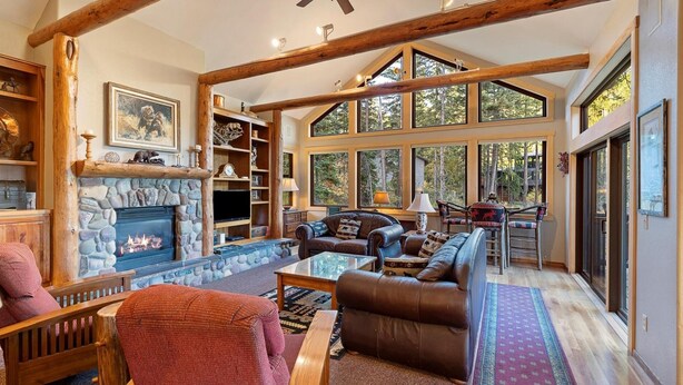 A spacious living room with a stone fireplace, leather sofas, and large windows offering views of the outdoors. - Rustic Ski-In/Ski-Out Townhome- Private Hot Tub, Game Room, Heated 2 Car Garage (Whitefish)