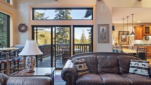 A spacious living room with a leather sofa, a dining area, and a large sliding glass door leading to a deck with outdoor seating. - Rustic Ski-In/Ski-Out Townhome- Private Hot Tub, Game Room, Heated 2 Car Garage (Whitefish)