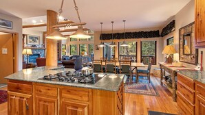 A spacious kitchen with granite countertops, wooden cabinets, and a central island with a stove. There is a dining area with a large table and chairs, and a cozy seating area with a fireplace. - Rustic Ski-In/Ski-Out Townhome- Private Hot Tub, Game Room, Heated 2 Car Garage (Whitefish)