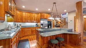 A kitchen with wooden cabinets, a stainless steel refrigerator, a granite countertop, and a bar stool. - Rustic Ski-In/Ski-Out Townhome- Private Hot Tub, Game Room, Heated 2 Car Garage (Whitefish)
