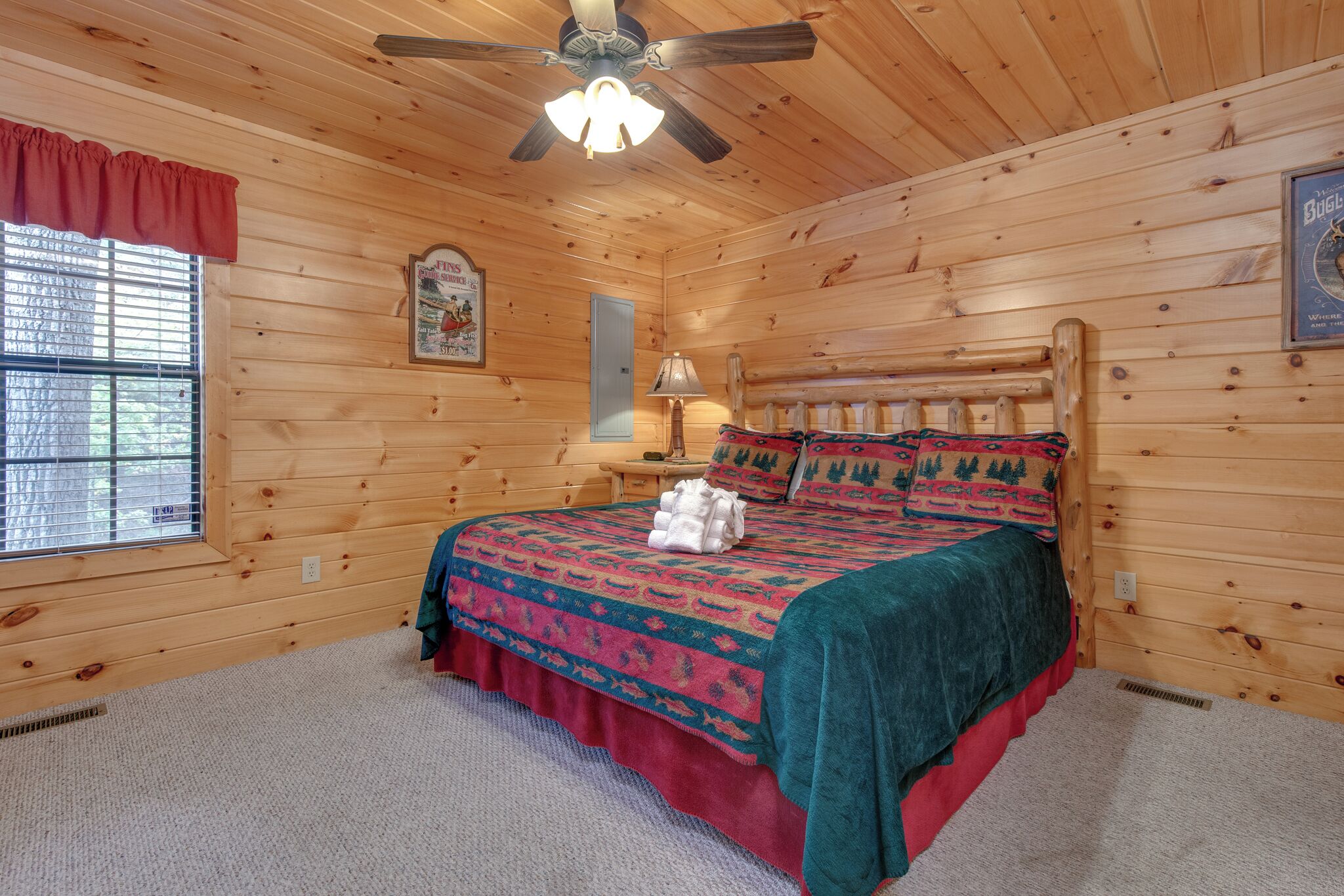 A wooden cabin bedroom with a bed, a ceiling fan, a window with blinds, and a framed poster on the wall.