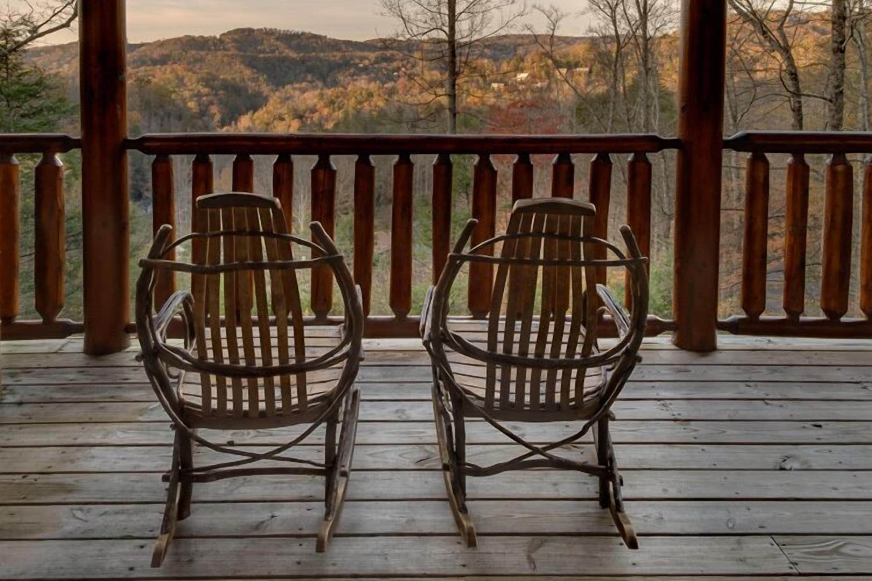 Two Adirondack chairs on a wooden deck with a railing, overlooking a mountainous landscape.