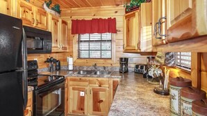 Une cuisine rustique aménagée dans une cabane en rondins, avec des meubles de rangement en bois, un réfrigérateur noir, une cuisinière, un four à micro-ondes, un évier et une fenêtre agrémentée d’un lambrequin rouge.