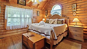 A log cabin bedroom with a wooden bed, a nightstand, a desk, and a window with a view. - Alpine Ski Lodge by Jackson Mountain Rentals (Gatlinburg)