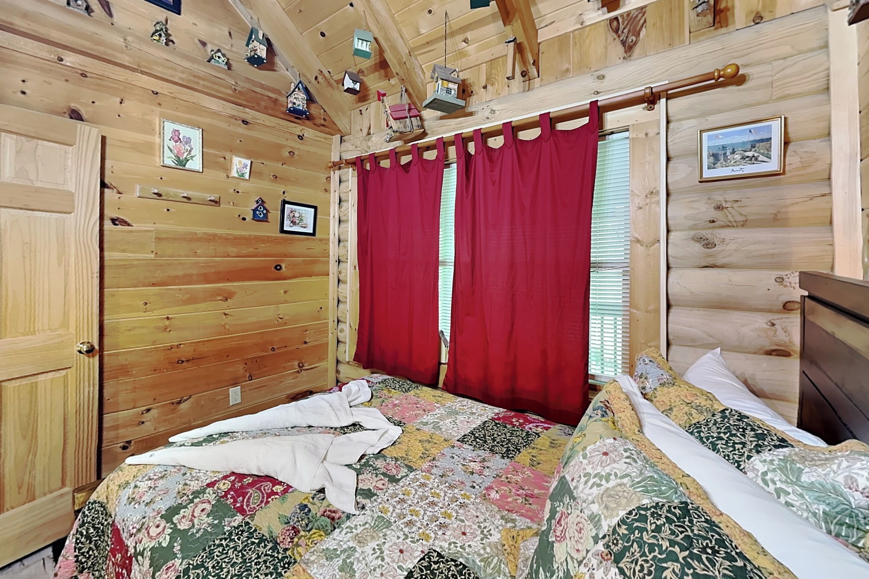 A wooden cabin bedroom with a bed, red curtains, and wall-mounted birdhouses.