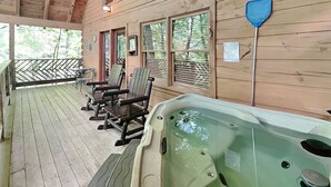 Una terraza de madera con mobiliario exterior, un jacuzzi y ventanas con vistas a una zona boscosa.