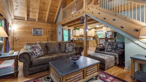 A spacious living room with a high ceiling, wooden beams, a staircase, and a view into the kitchen area. - Above Ole Smoky's Clouds by Jackson Mountain Rentals (Gatlinburg)