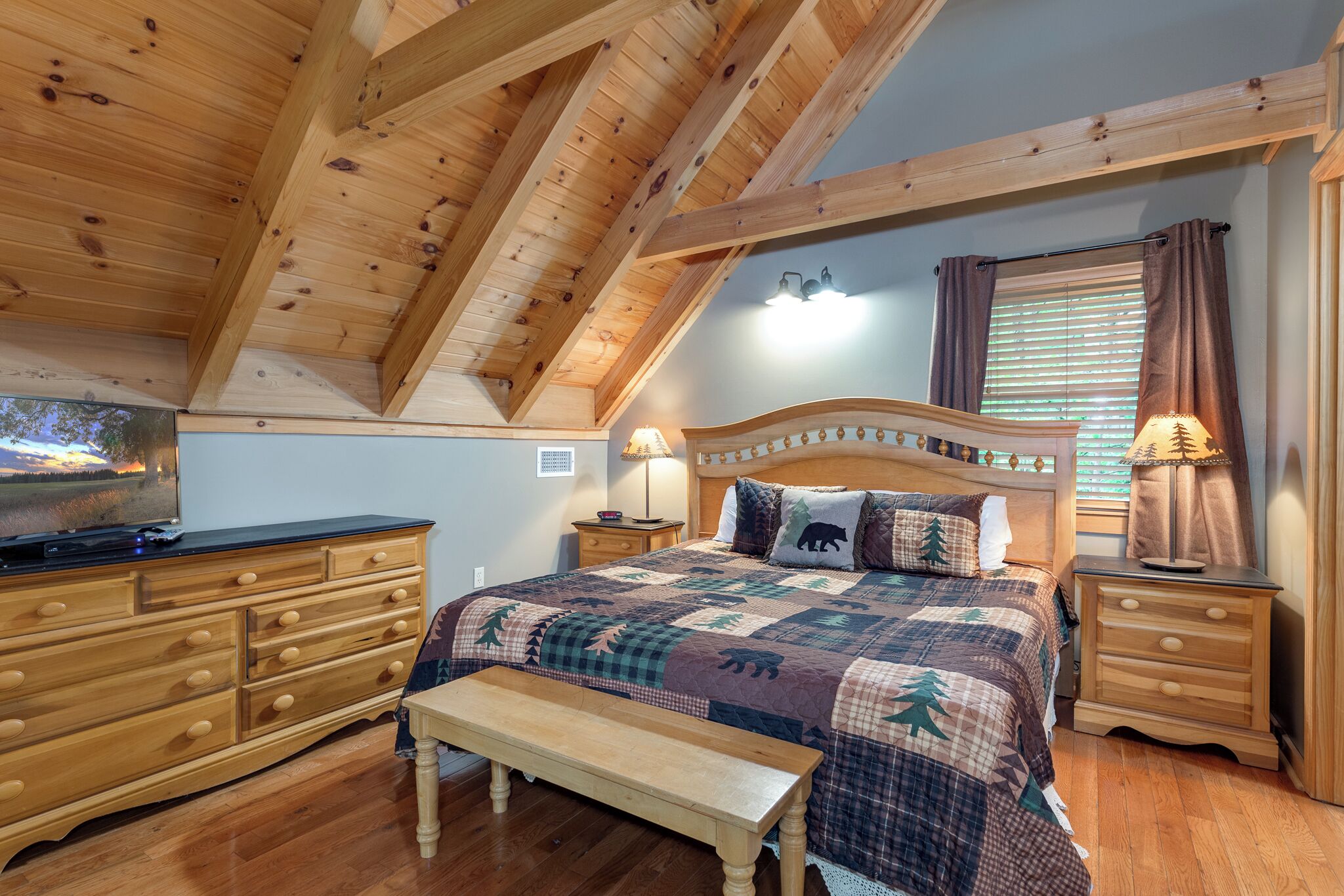 A bedroom with a wooden ceiling, a bed with a quilt, a bench, a dresser, and a nightstand with lamps.