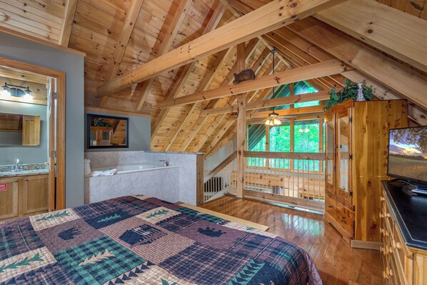 A cozy attic bedroom with a wooden ceiling, a large bed, a built-in TV, and a view of the outdoors.