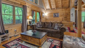 Interior acogedor de una cabaña con techo de madera, un sofá de cuero, una mesa de centro y vistas al bosque a través de amplios ventanales.