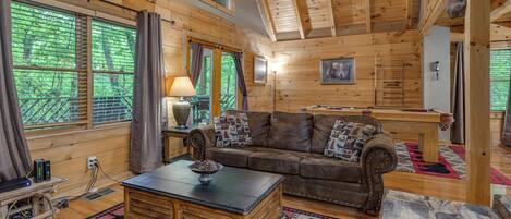 Interior acogedor de una cabaña con techo de madera, un sofá de cuero, una mesa de centro y vistas al bosque a través de amplios ventanales.