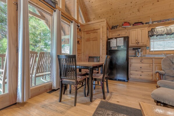 L’intérieur d’un chalet en bois comprend une salle à manger avec une table et des chaises, un coin cuisine avec un réfrigérateur, et de grandes fenêtres dotées de rideaux.