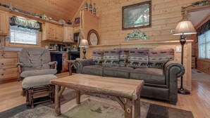 A cozy cabin interior with a wooden dining table, a cushioned armchair, a sofa with patterned cushions, and a kitchen area with wooden cabinets and a hanging lamp. - Americana by Jackson Mountain Rentals (Gatlinburg)
