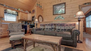 Interior acolhedor de cabana com mesa de jantar de madeira, poltrona confortável, sofá com almofadas estampadas e cozinha com armários de madeira e luminária pendente.