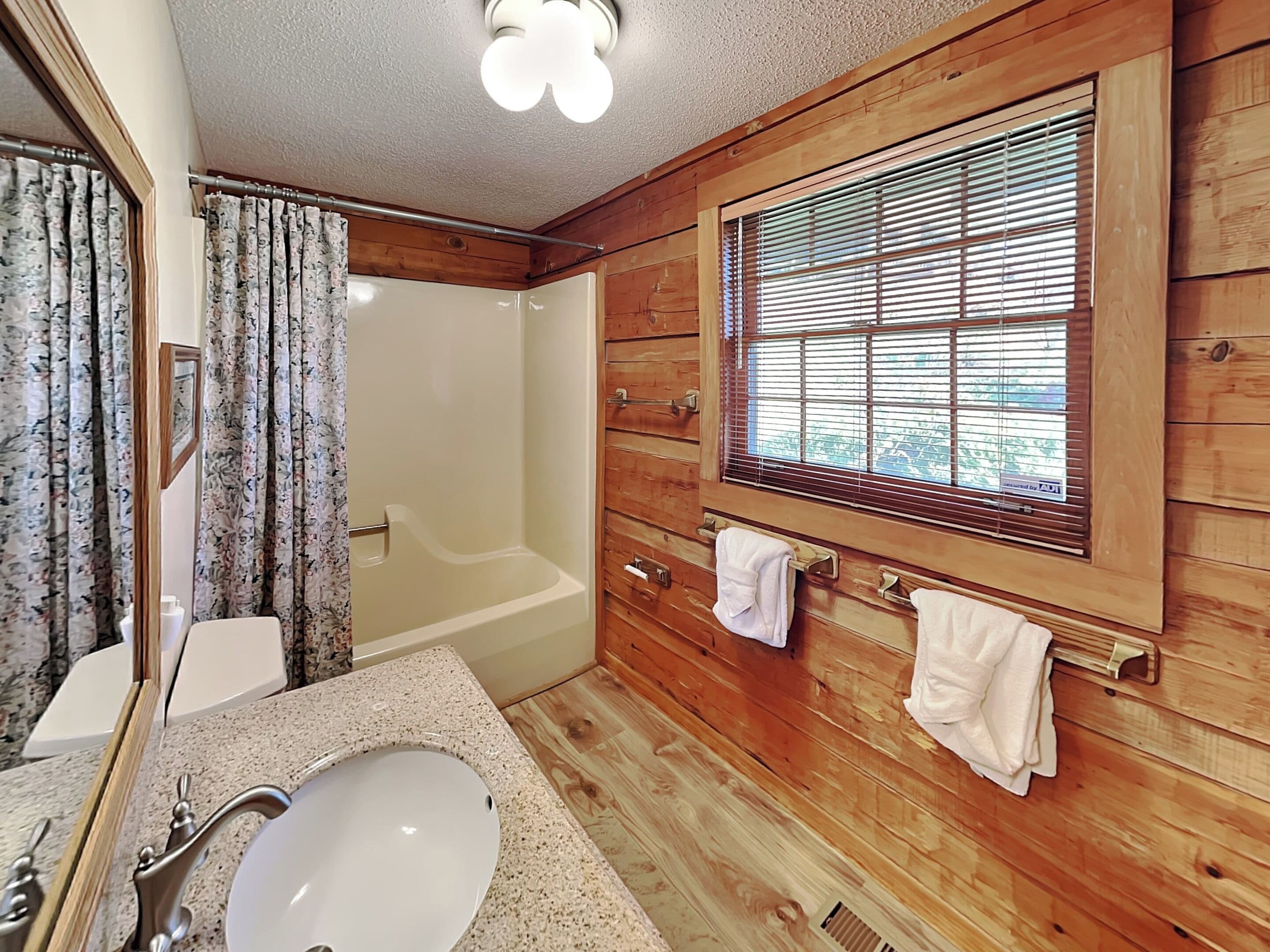 A wooden cabin bathroom with a bathtub, a sink, and a window with blinds.