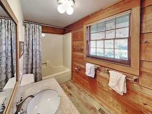 A wooden cabin bathroom with a bathtub, a sink, and a window with blinds.