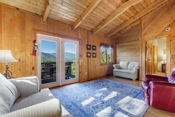 A wooden cabin interior with a sofa, a red chair, a blue patterned rug, and a view through the door.