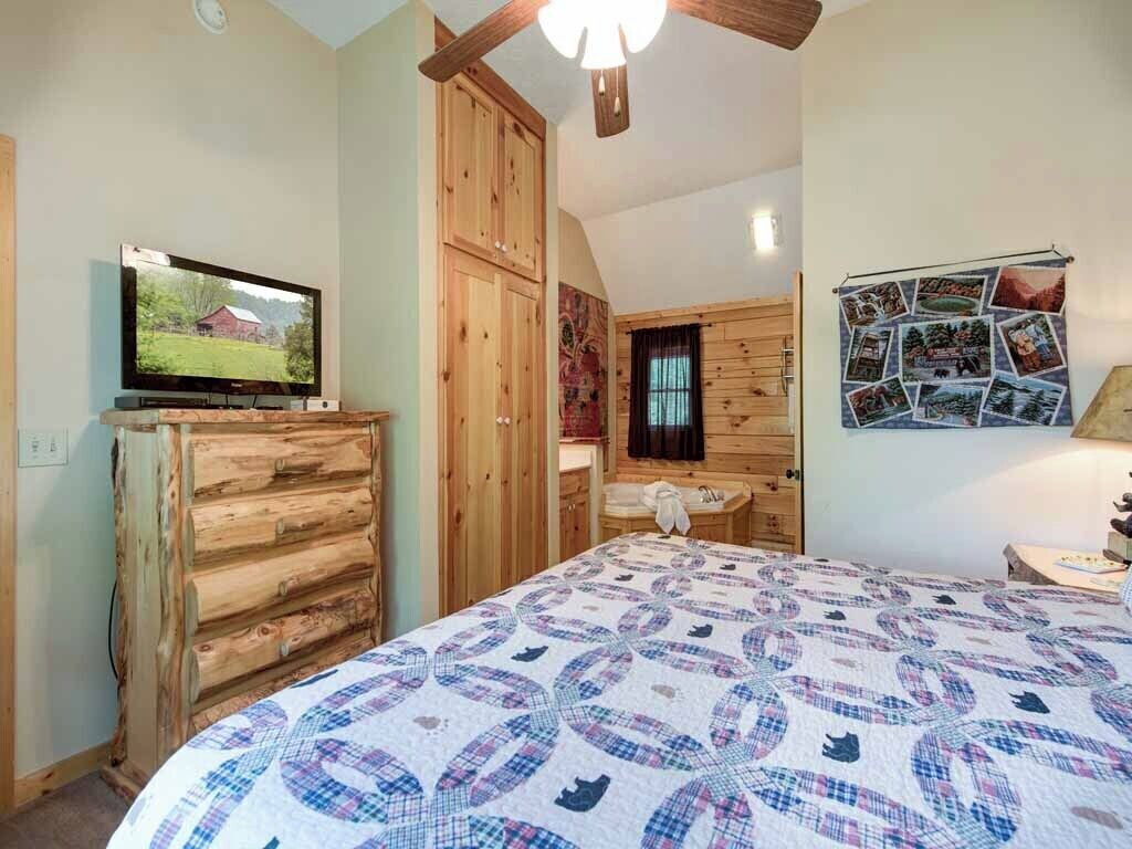 Une chambre avec une commode en bois, un lit recouvert d’une courtepointe, un téléviseur posé sur une étagère et un mur orné de photos.