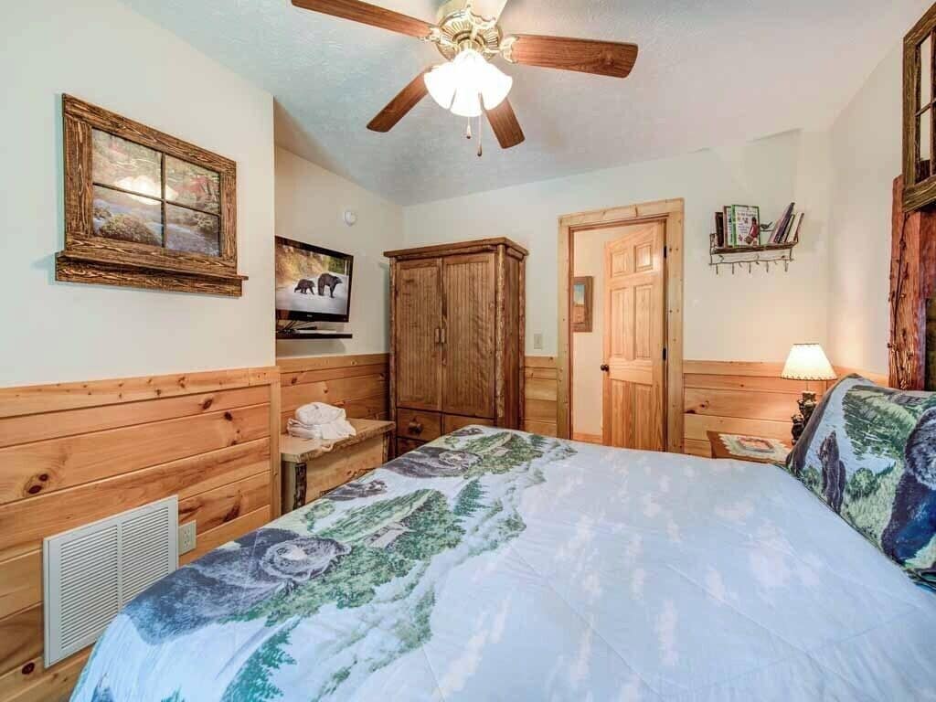 A bedroom with wooden paneling, a bed with a floral bedspread, a ceiling fan, a television, and a wooden wardrobe.