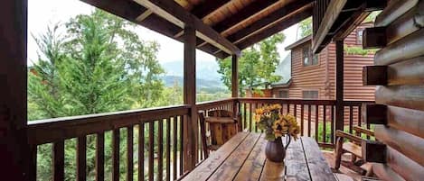 A wooden deck with a table and chairs, a vase of flowers, and a view of a forested mountain area.