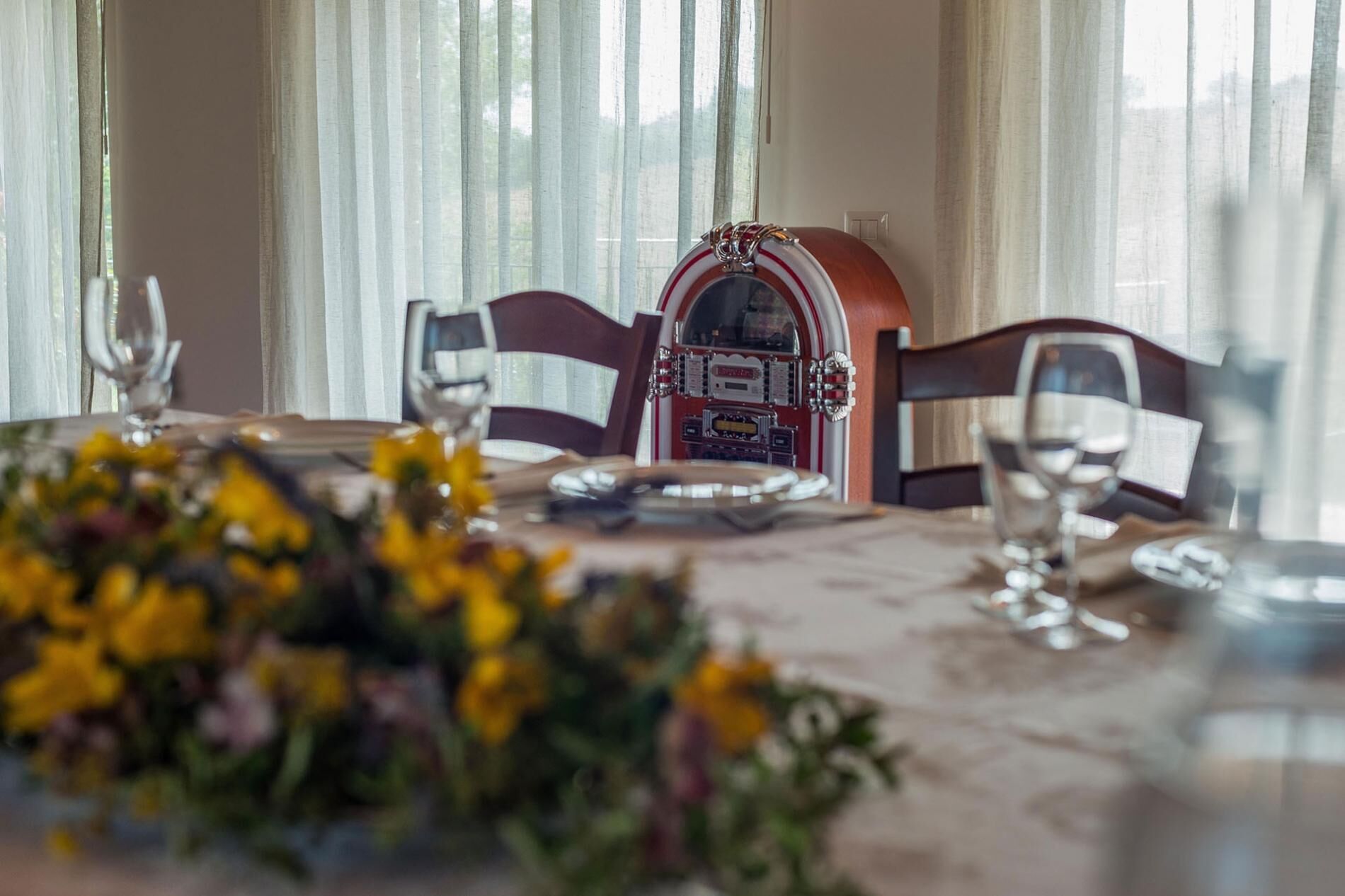 Una mesa de comedor dispuesta con un florero de flores amarillas, un jukebox y una ventana con cortinas.